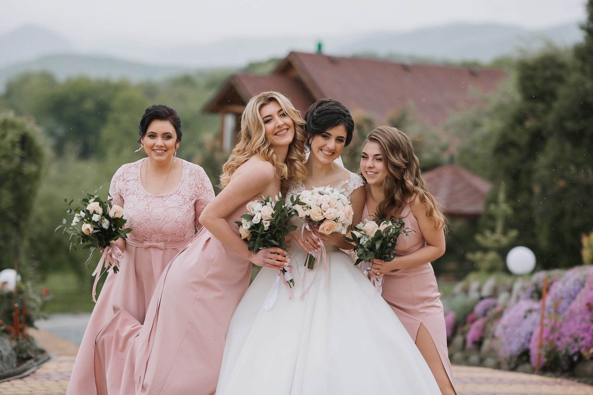 Bride and bridesmaids in pink dresses pose outdoors, holding bouquets.