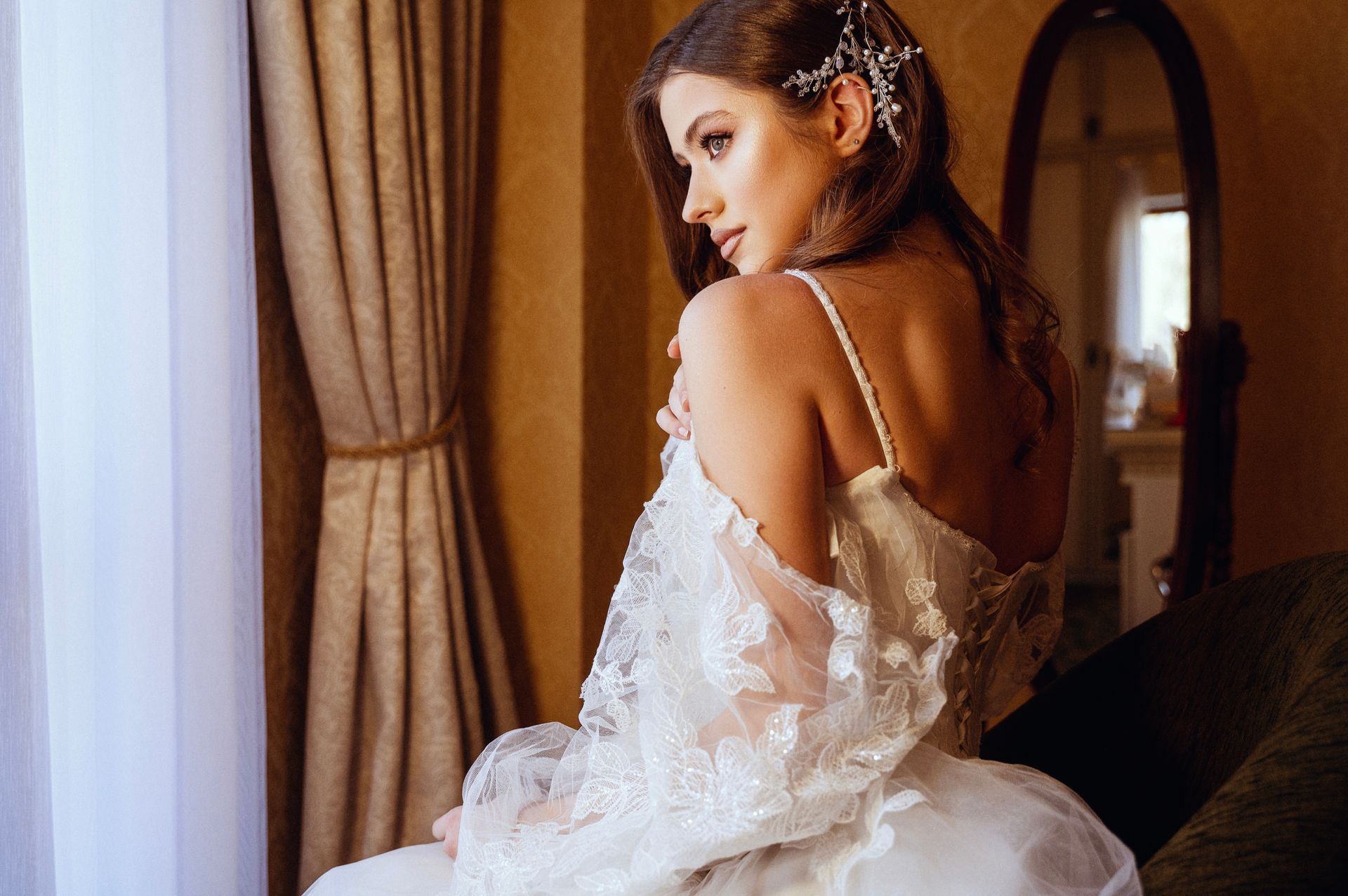 Woman in white lacy dress looking over her shoulder by window, hand on dress, soft lighting.