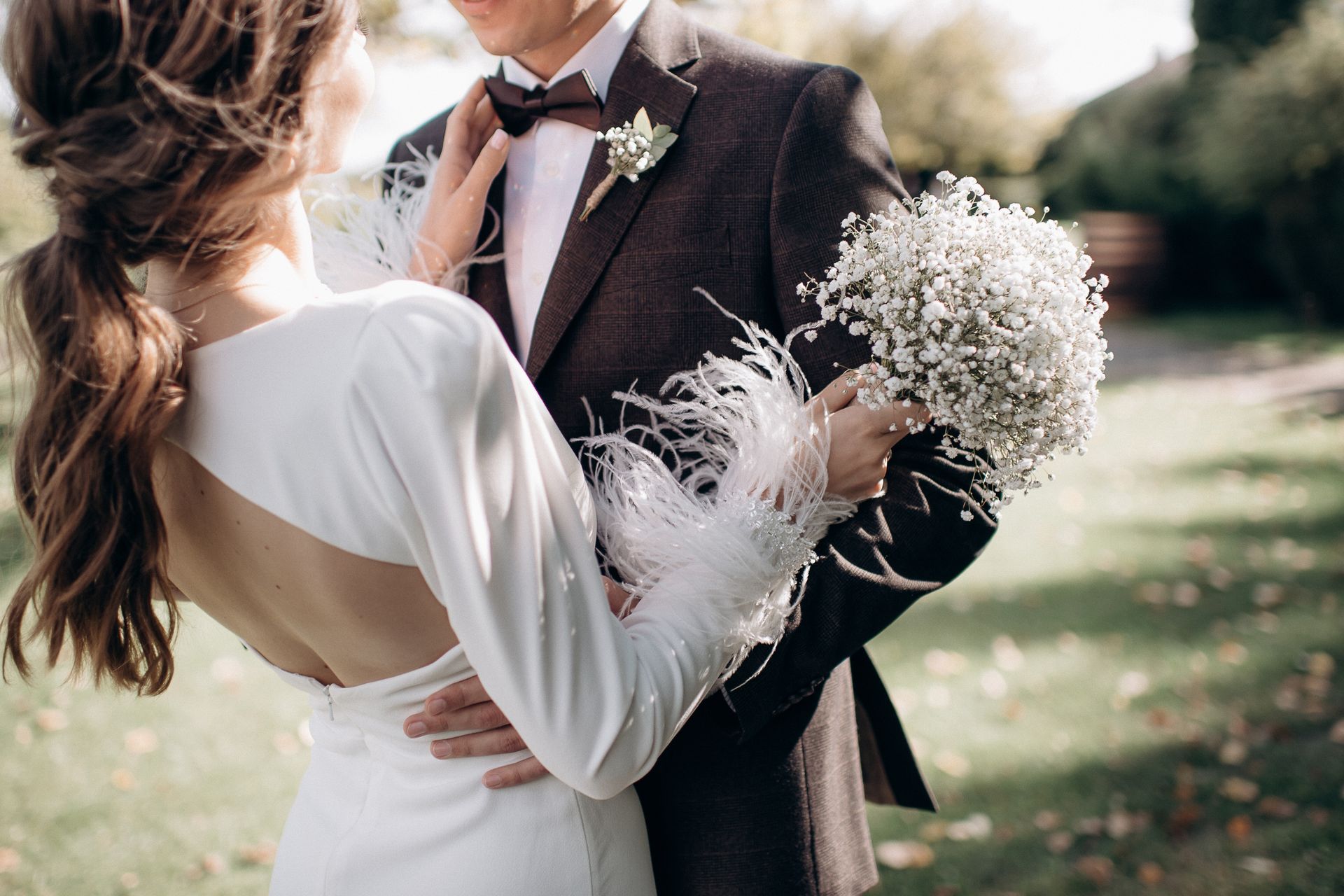Bride in white dress with feathered sleeves embraces groom in a suit, holding baby's breath bouquet. Outdoor setting.