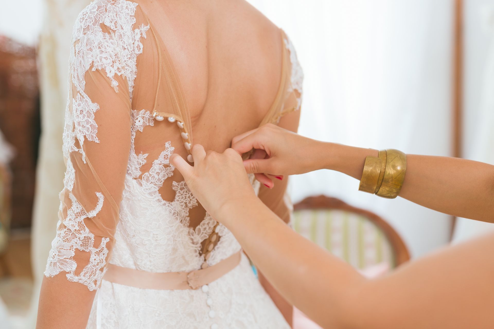 Woman being buttoned into a white lace wedding dress; a helping hand with gold bracelets.