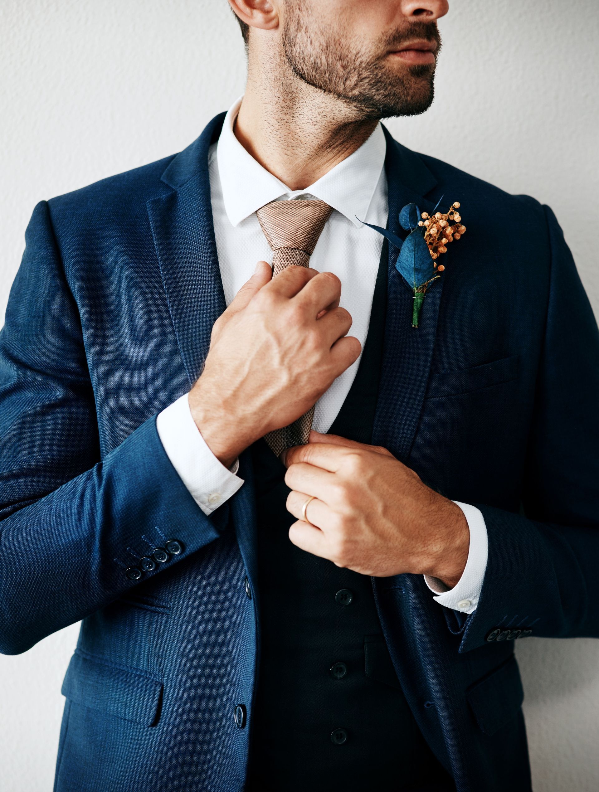 Man in blue suit adjusting tie, white shirt, and boutonniere.