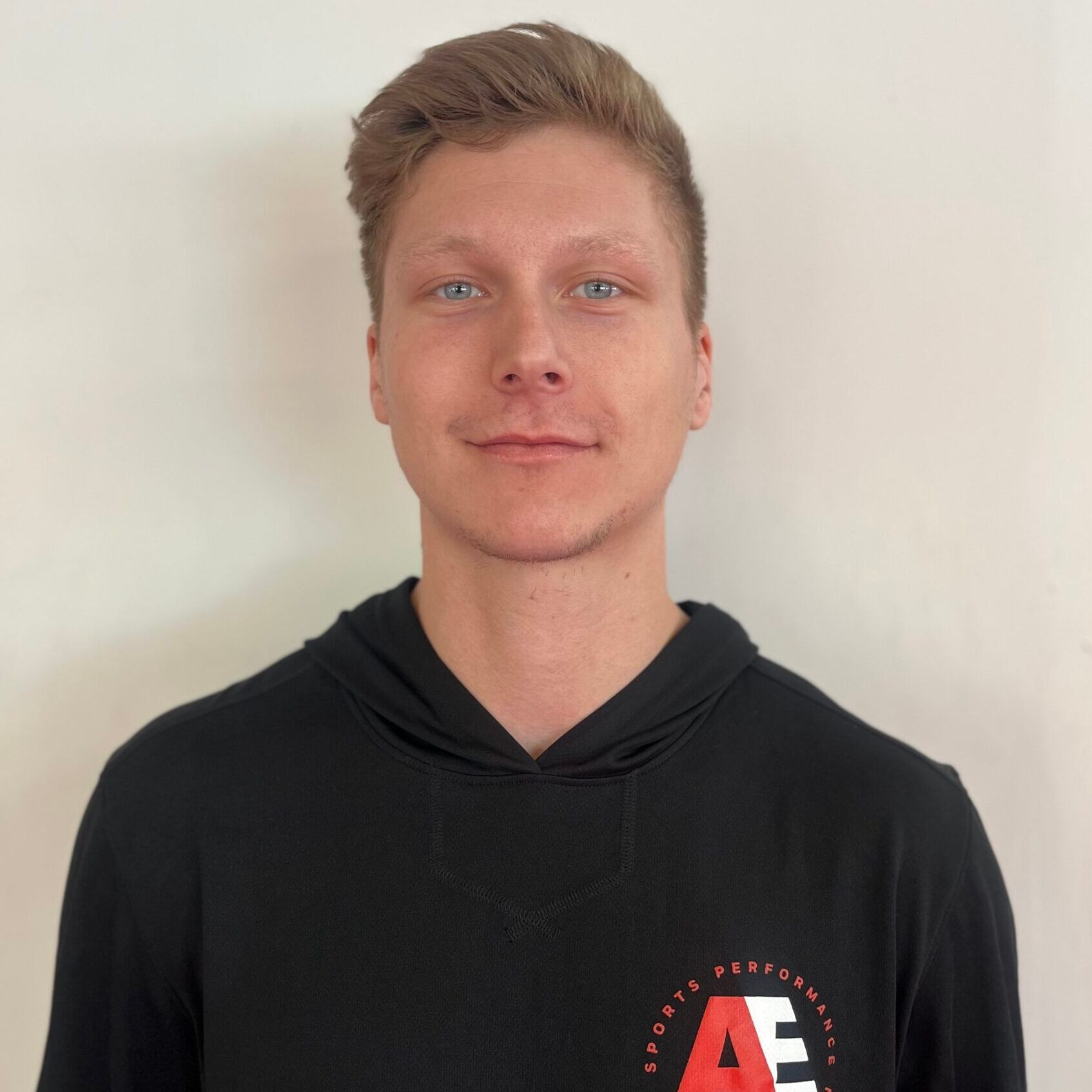 A young man wearing a black hoodie is standing in front of a white wall.