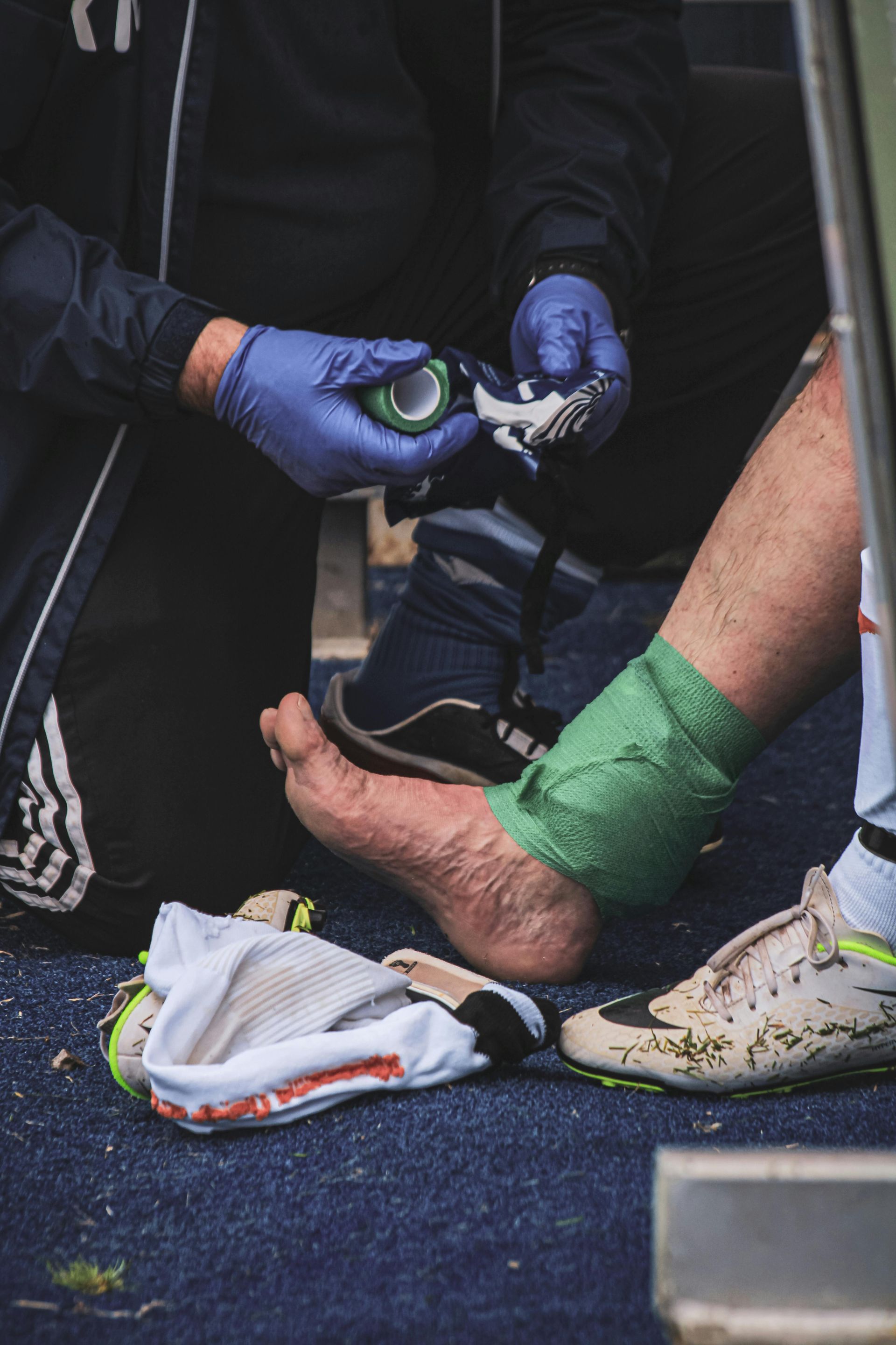 Person's ankle being bandaged after an injury. Person is wearing a soccer cleat, in an outdoor setting.