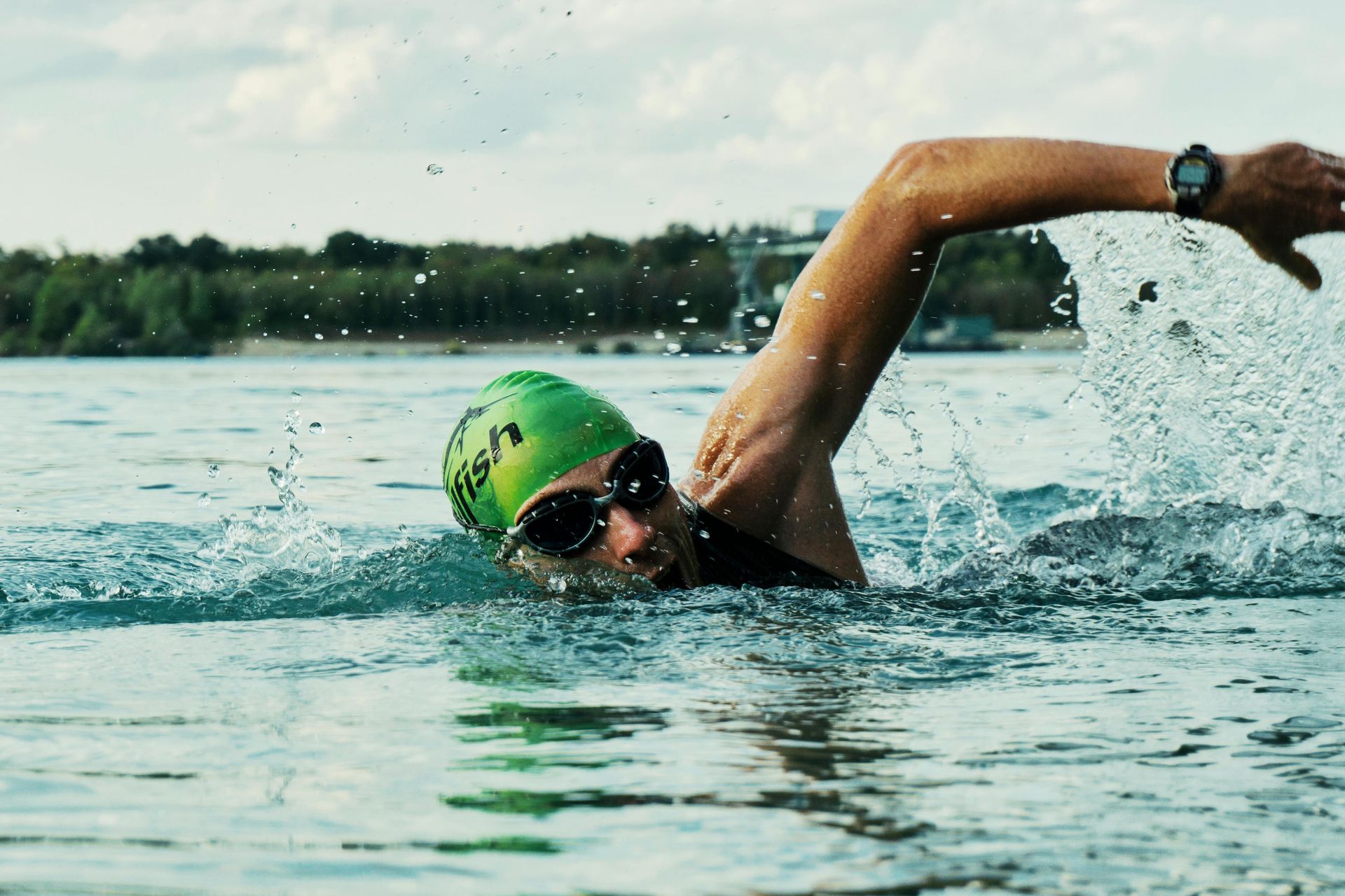 Swimmer in a green cap and sunglasses, in open water, arm outstretched, water splashing.