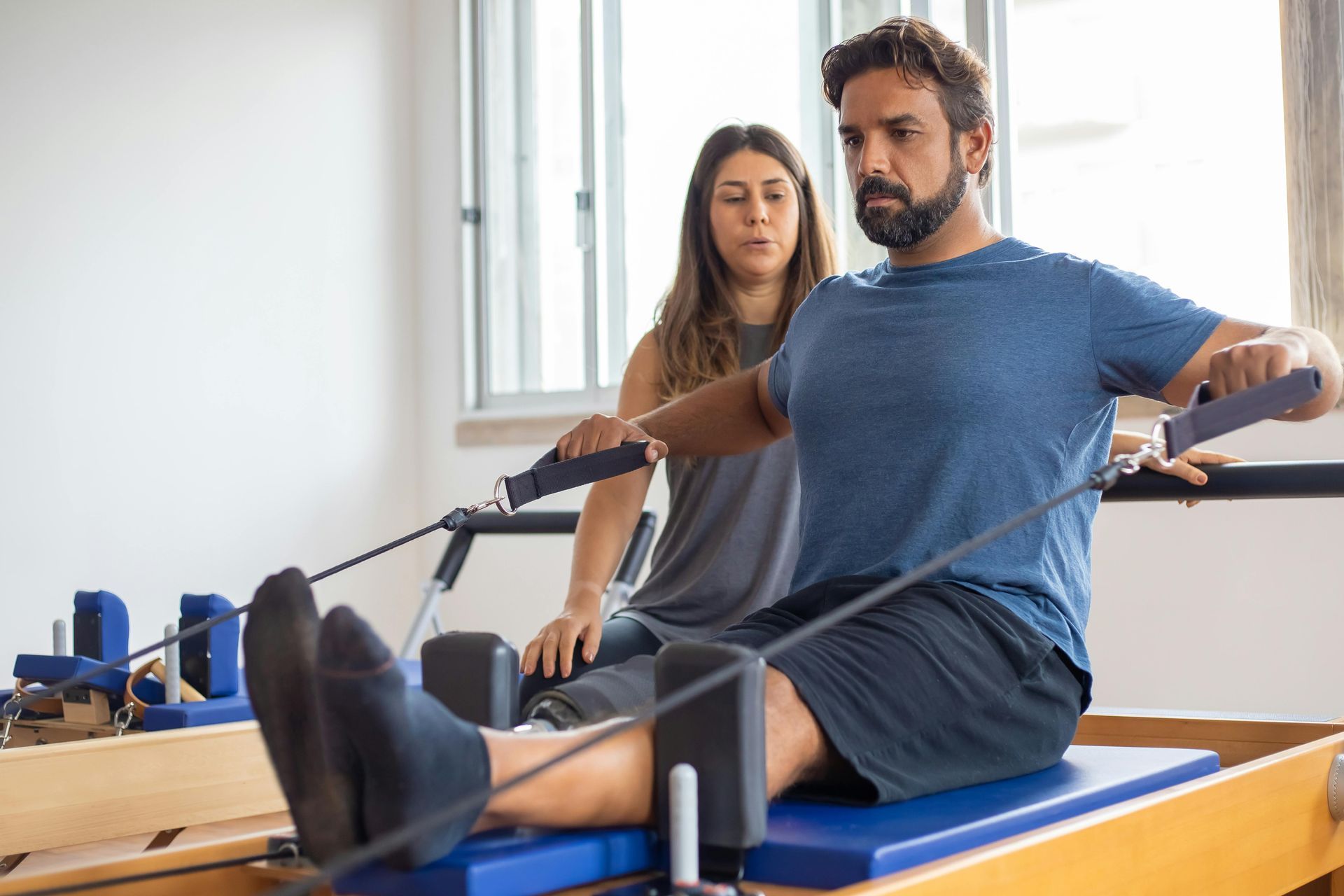 Man doing Pilates exercise with instructor; seated, pulling handles.
