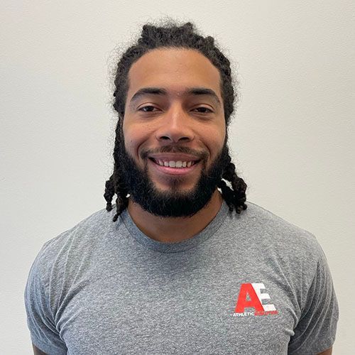 A man with dreadlocks and a beard is wearing a grey shirt and smiling.