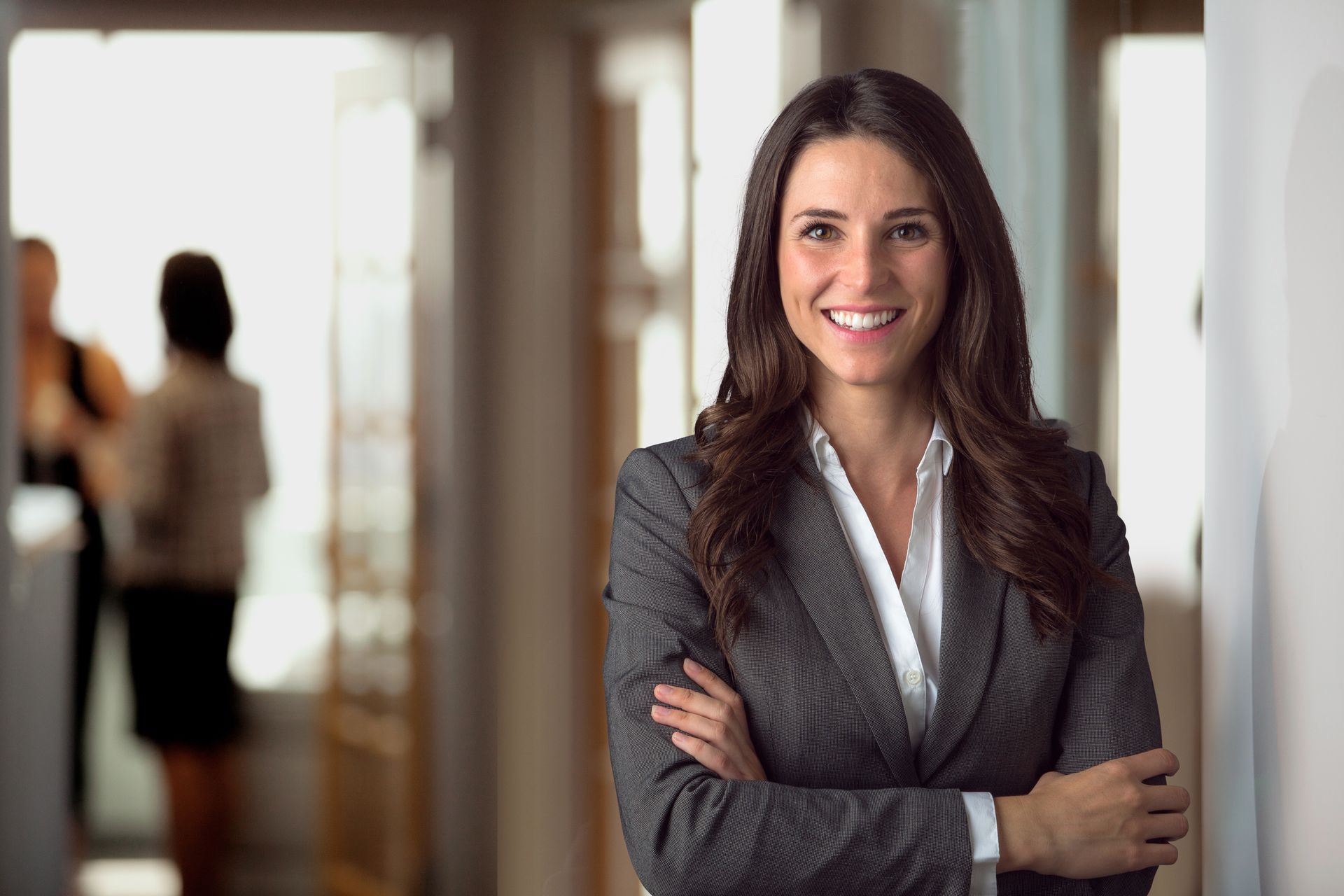 A woman in a suit is standing in a hallway with her arms crossed and smiling.