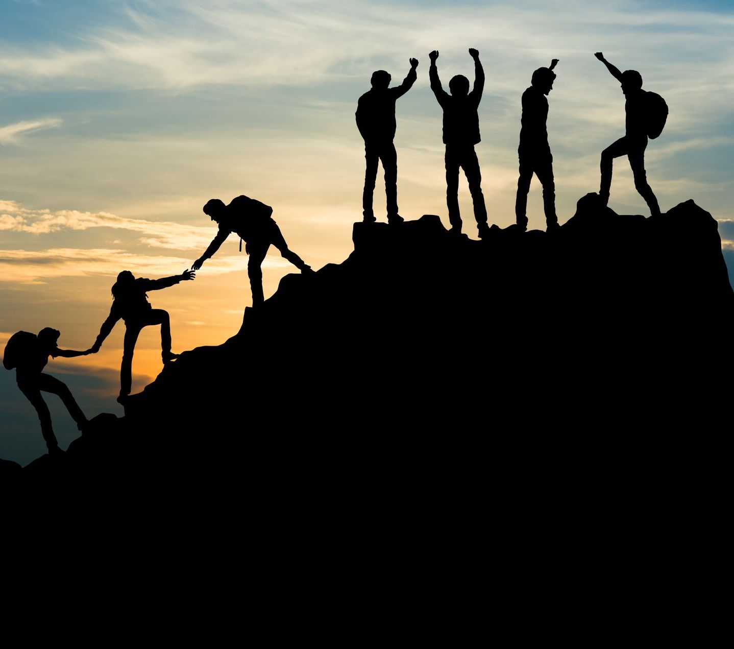 A silhouette of a team helping each other making it up a mountain while the sun sets in the background. The people at the top are celebrating.