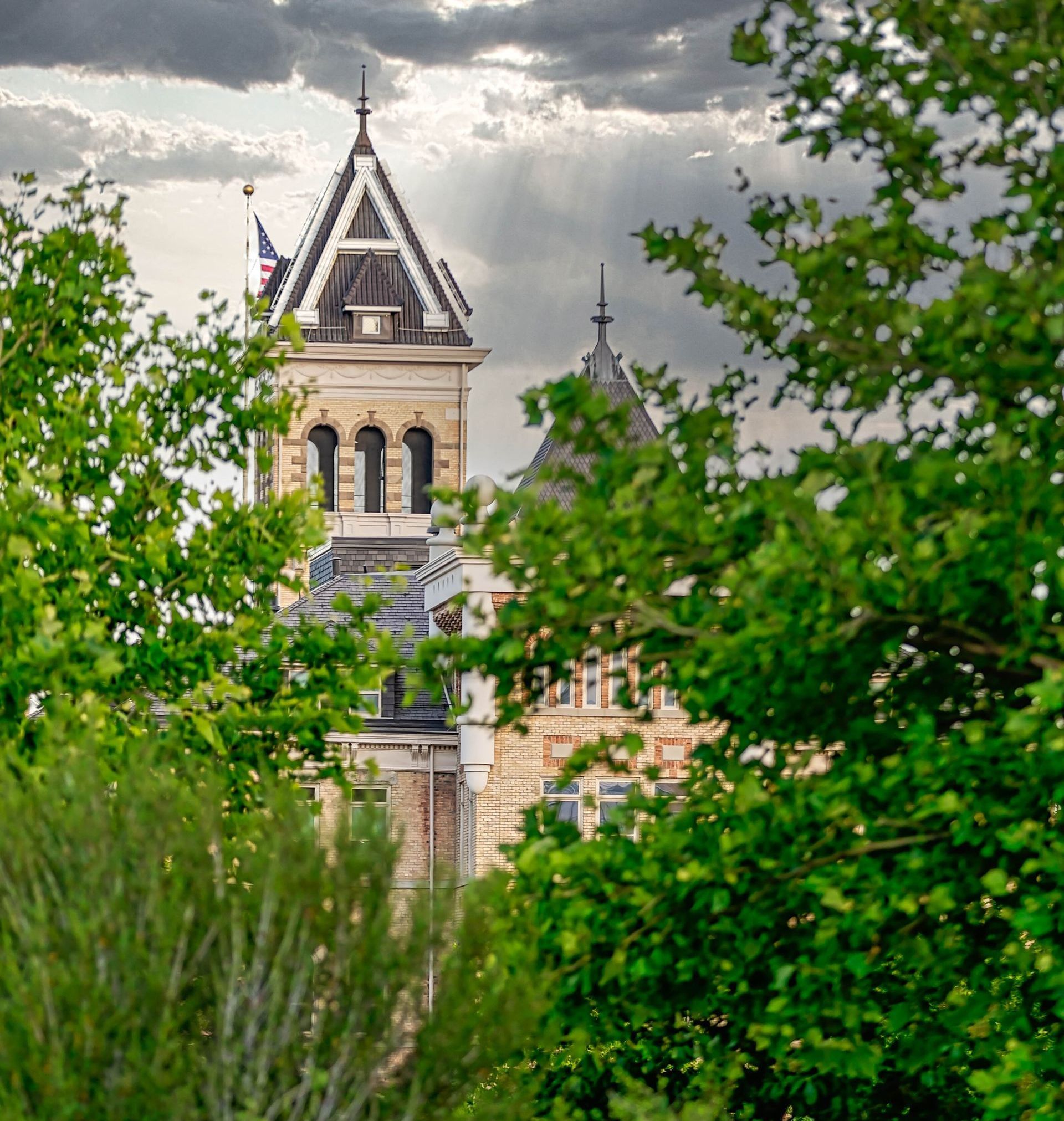 An image of Utah State University's Old Main building with green trees in the foreground and a cloudy sky.