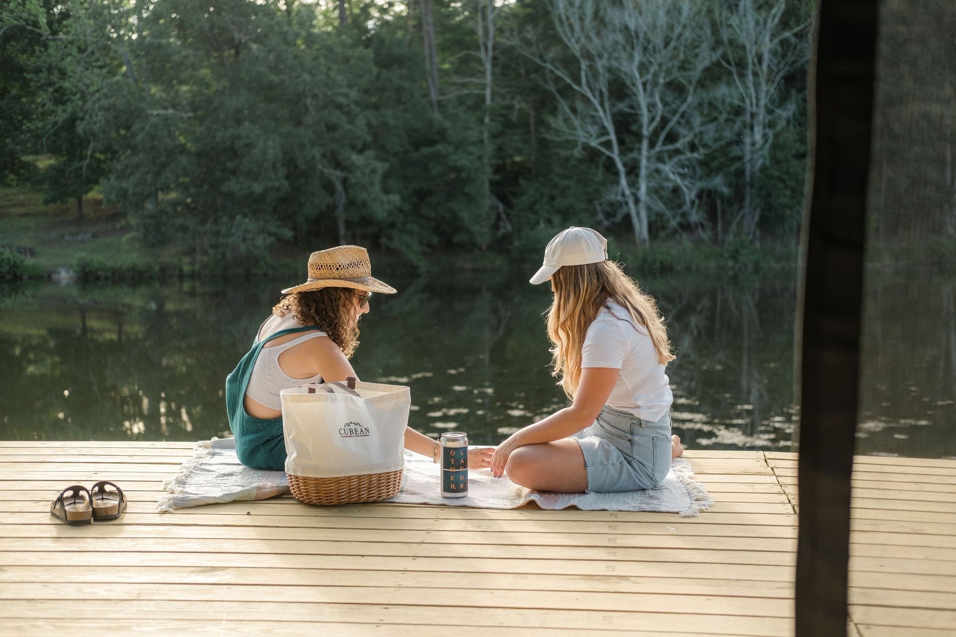 Two women are sitting on a dock near a lake having a picnic.