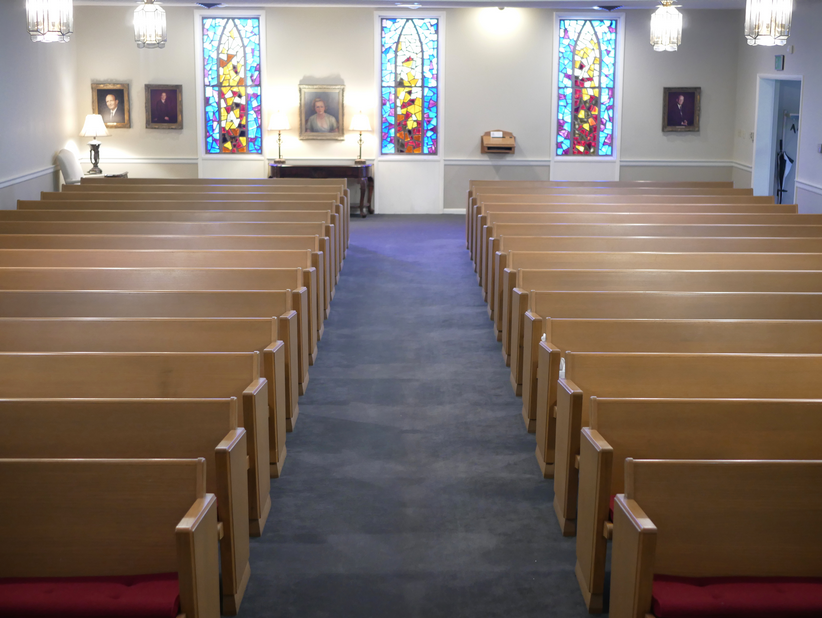 Interior of a chapel with rows of wooden pews, stained-glass windows, and portraits on the walls.
