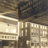 Old sepia photograph of a building with a Roberts Funeral Home sign. Other buildings in the background.