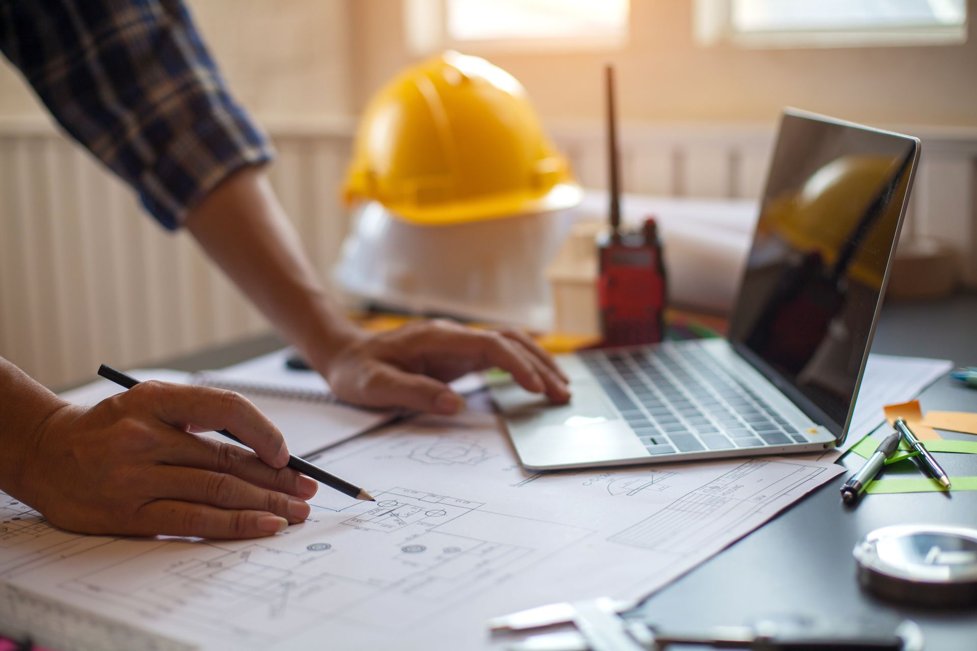 Person working on blueprints at a desk with a laptop, hard hats, and radio.