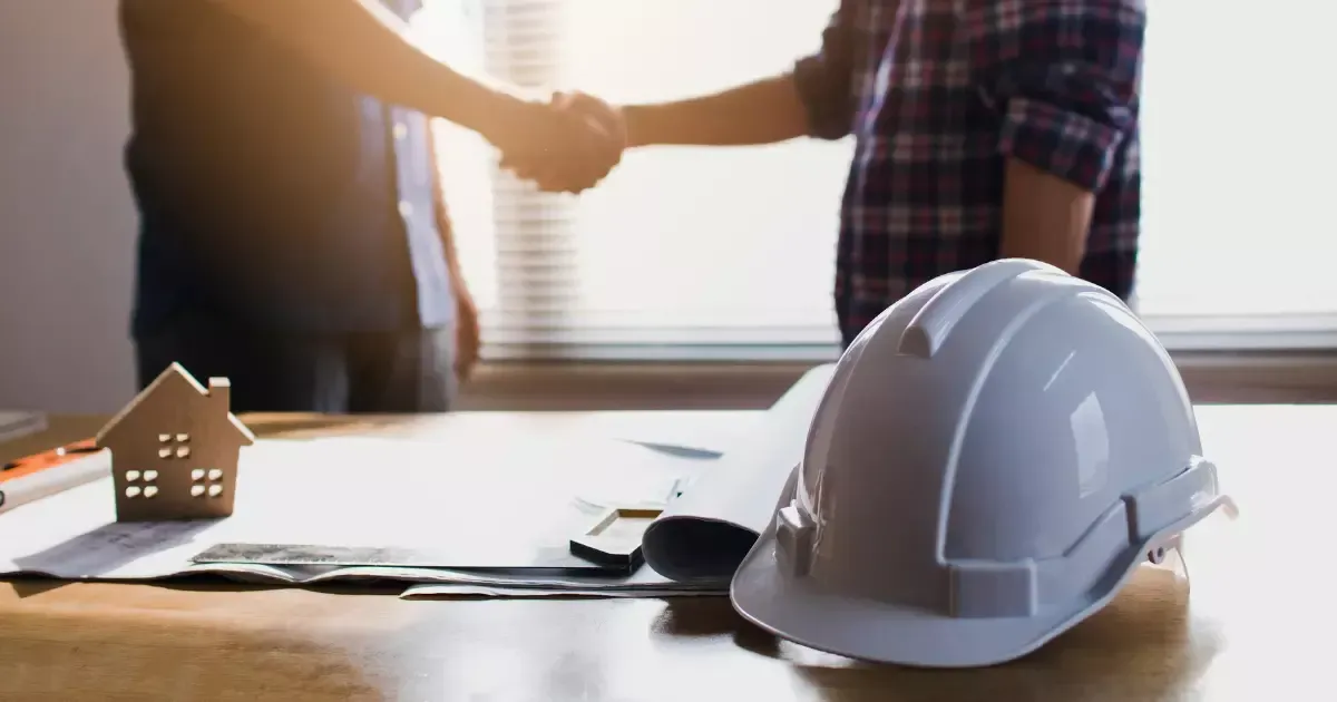 Builders shaking hands next to safety helmets and plans
