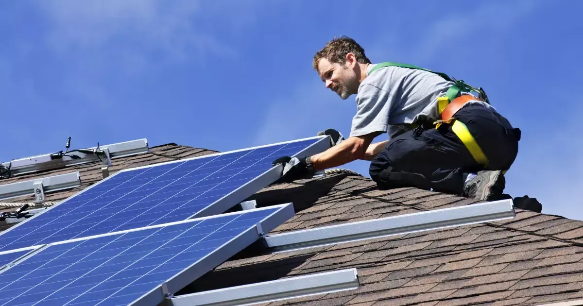 Solar panel installer securing a photovoltaic panel on a residential shingle roof under a clear sky
