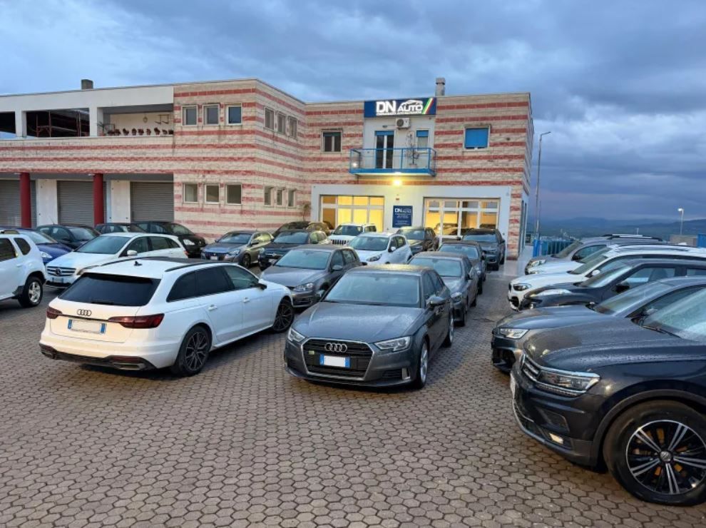 Un parcheggio pieno di auto di vario tipo davanti a un edificio commerciale in mattoni a più piani, sotto un cielo nuvoloso.