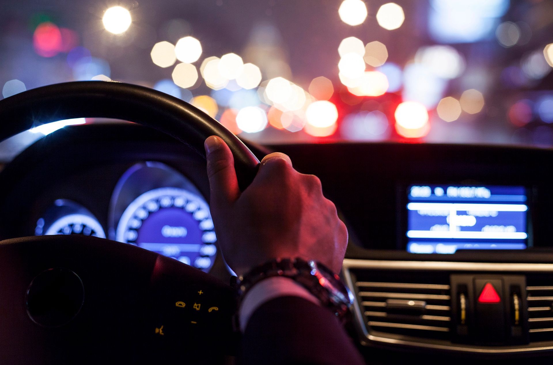 Di notte, le mani di un guidatore stringono il volante, con gli indicatori del cruscotto e i lampioni sfocati della città sullo sfondo.