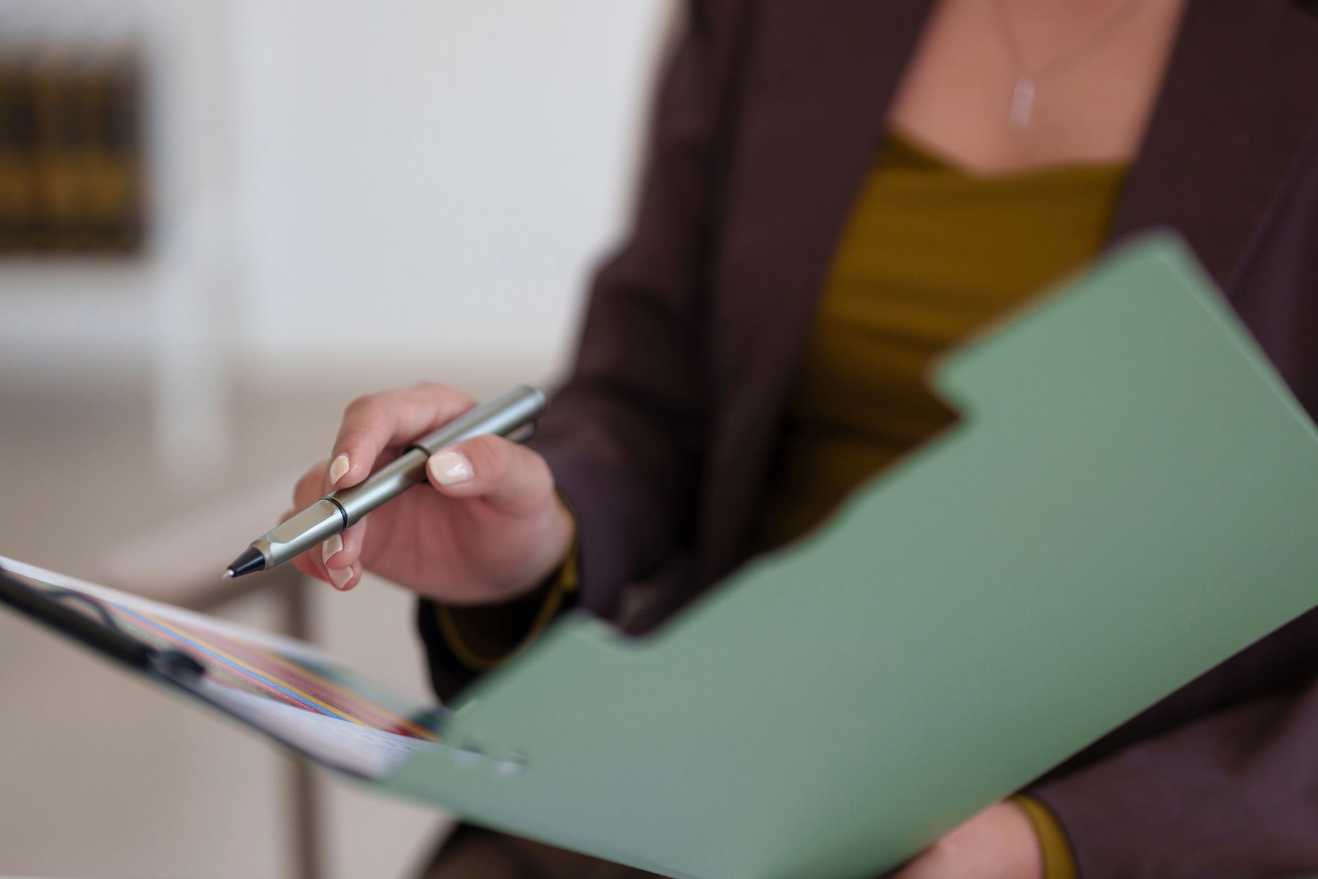 Una persona con un blazer marrone tiene in mano una cartella verde e una penna, indicando il documento aperto.