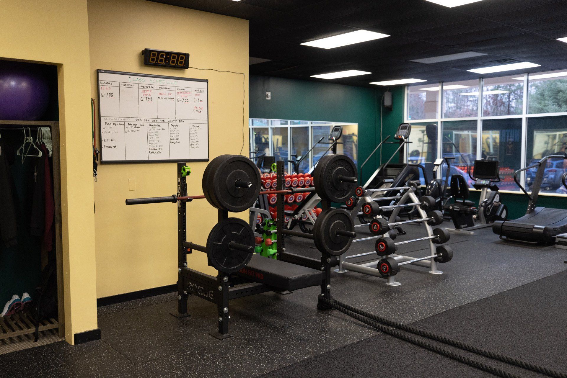 A small gym featuring a weight rack with loaded barbells, a whiteboard, workout machines, and exercise equipment.