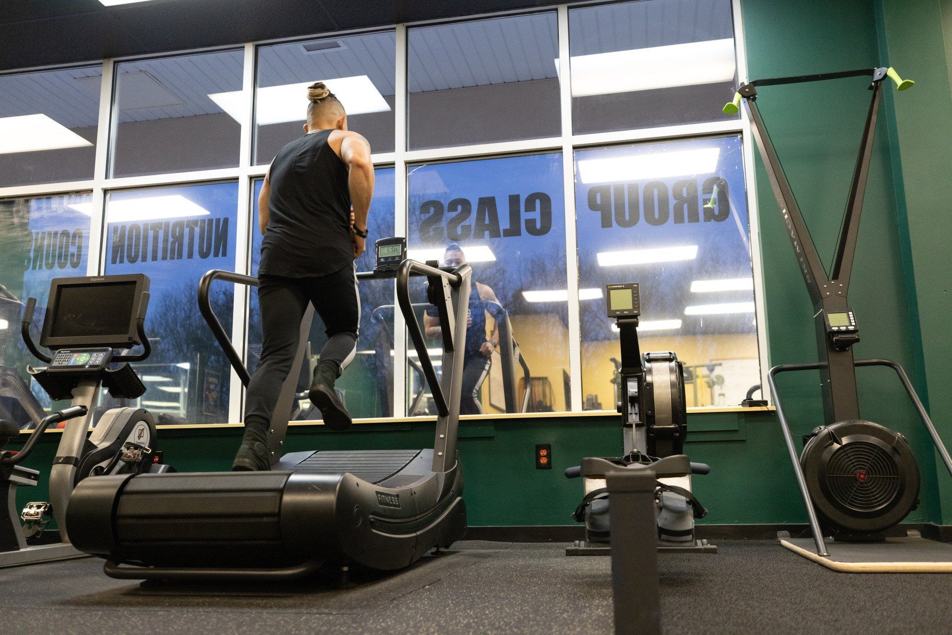 A person runs on a curved manual treadmill in a gym, with fitness equipment and mirrored windows in the background.