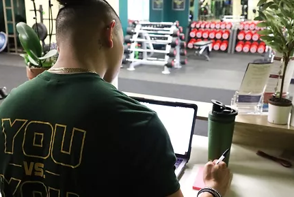 A person in a green shirt sits at a desk in a gym, writing in a notebook while looking at a laptop screen.