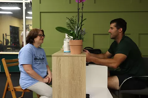 Two people sit at a desk in a room with green walls, facing each other while engaged in a conversation across a plant.