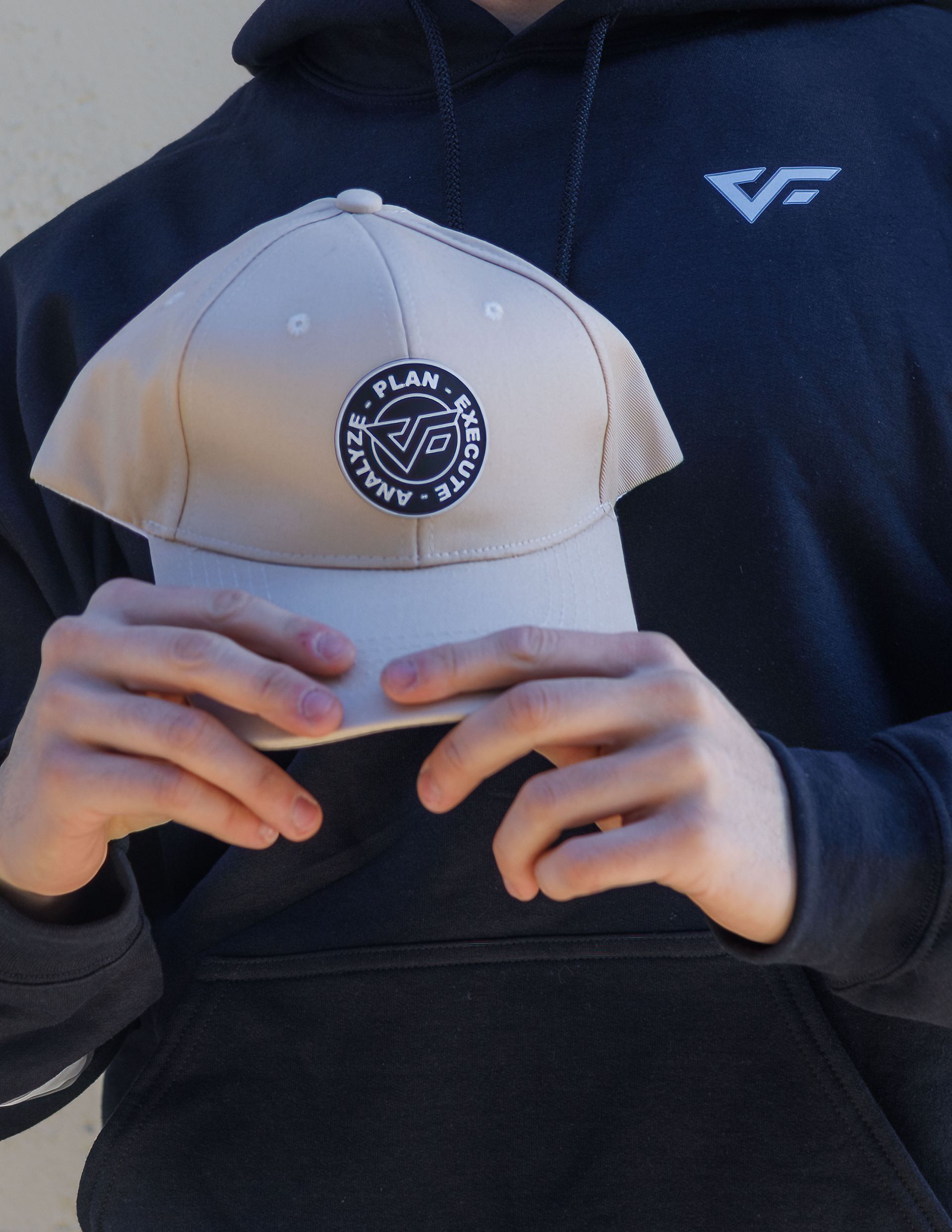 A person wearing a dark hoodie holds up a beige baseball cap featuring a circular logo with text and a geometric design.