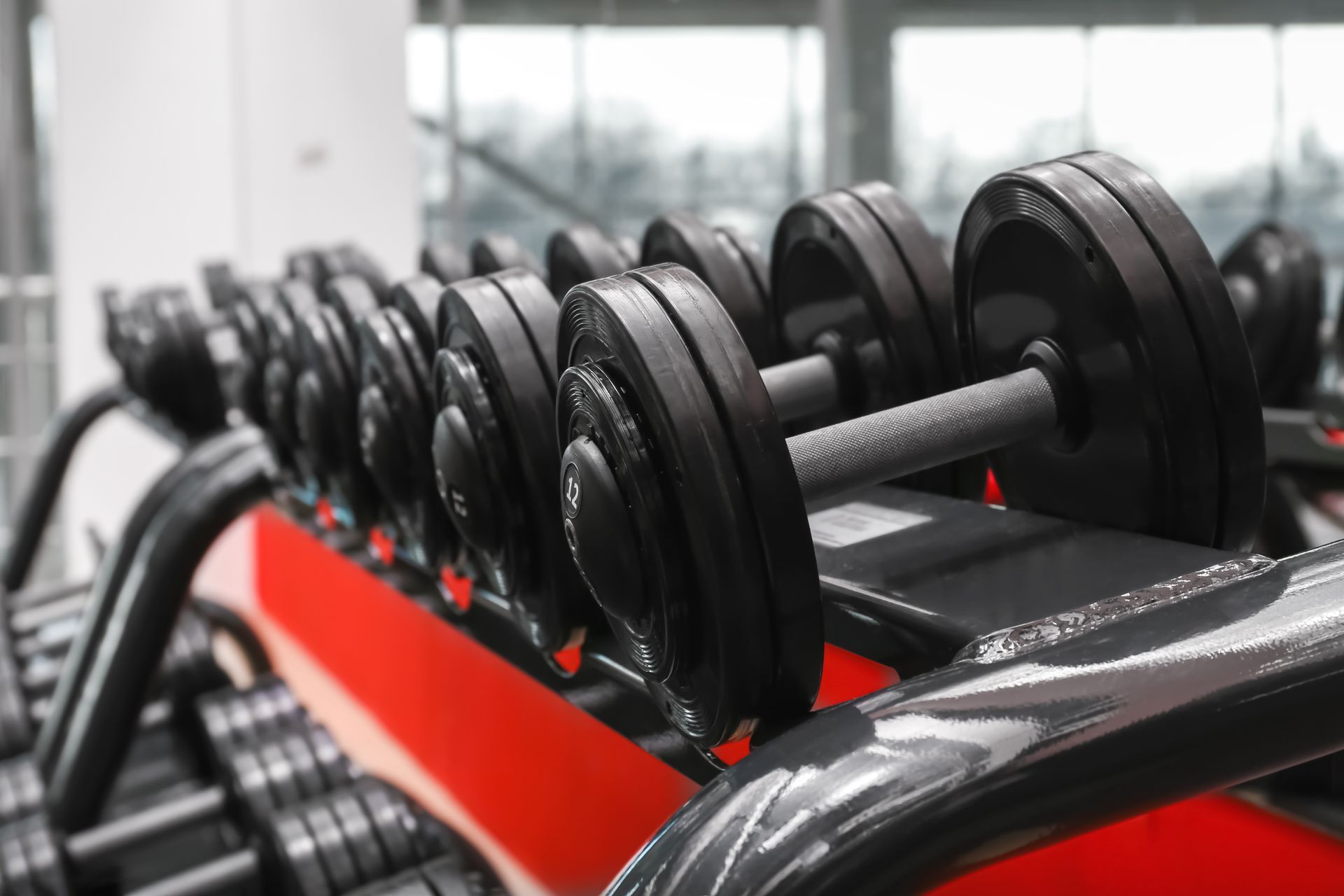 A row of black dumbbells on a rack in a gym, with the foreground weight in focus and the rest blurred.