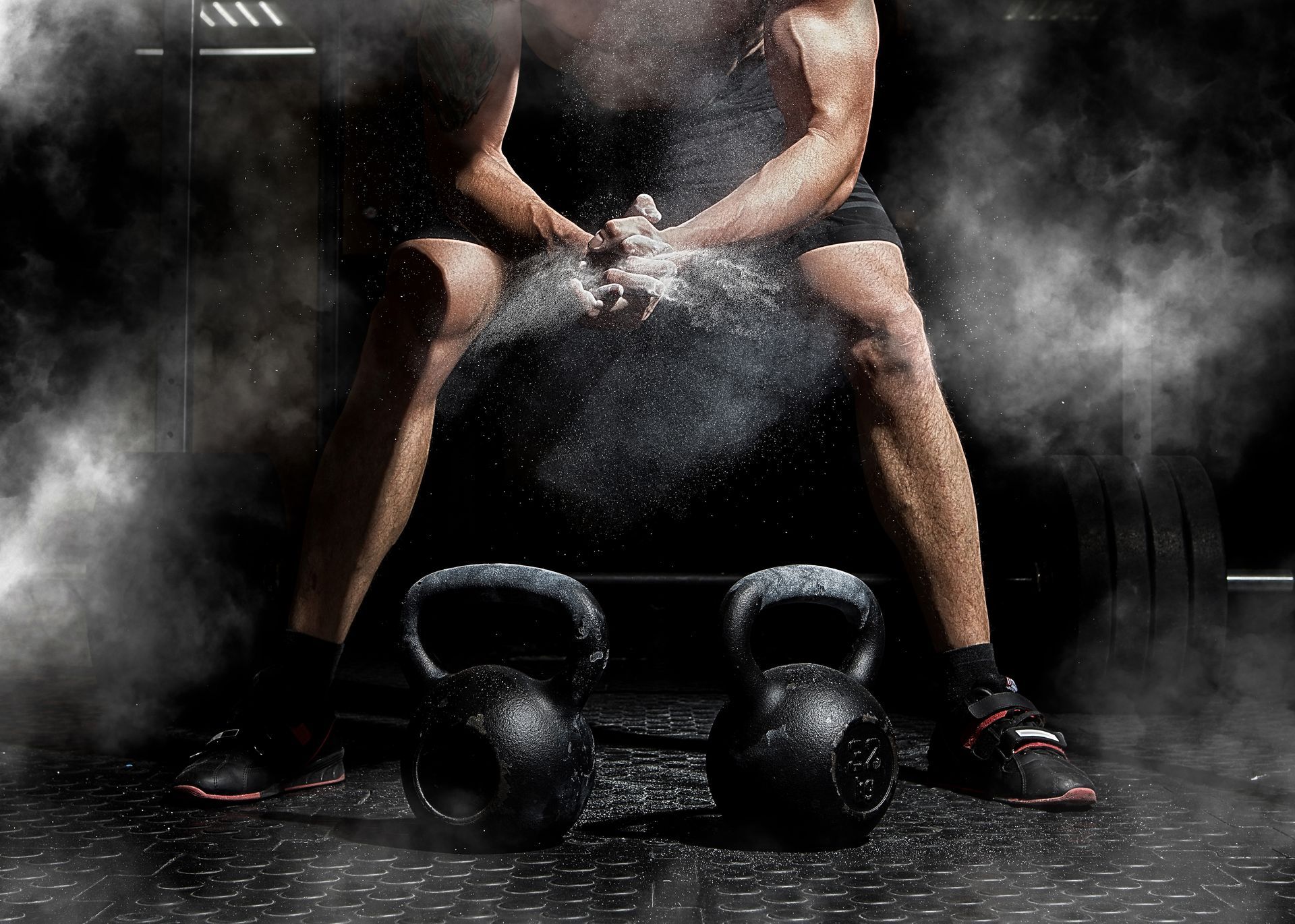 A person sits on a gym floor, clapping hands filled with chalk dust between two kettlebells, surrounded by smoke.