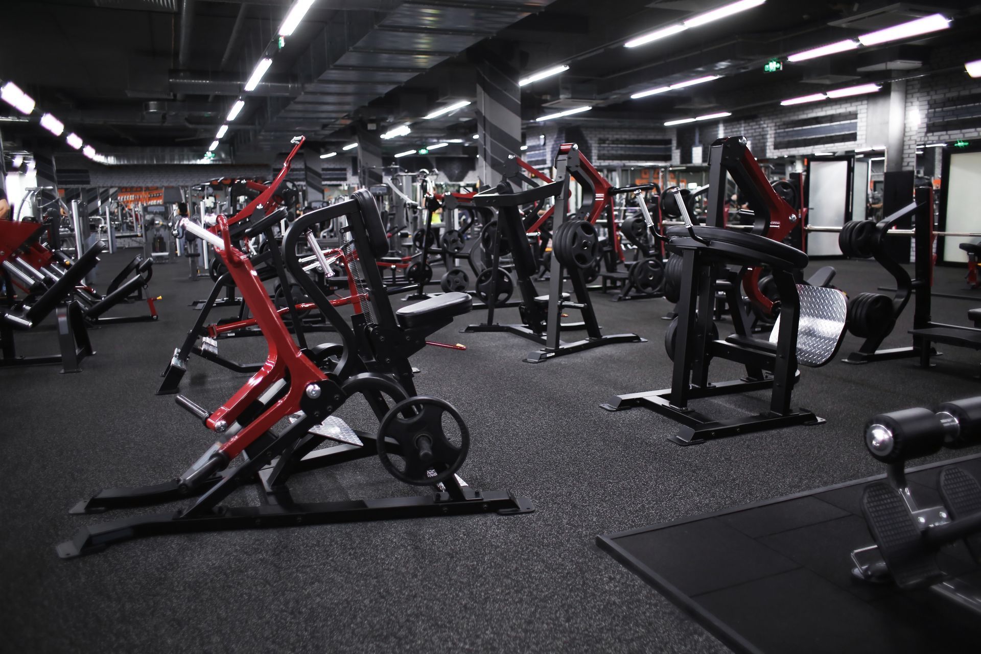 A gym floor filled with black and red weight machines and rubber flooring under bright overhead lights.