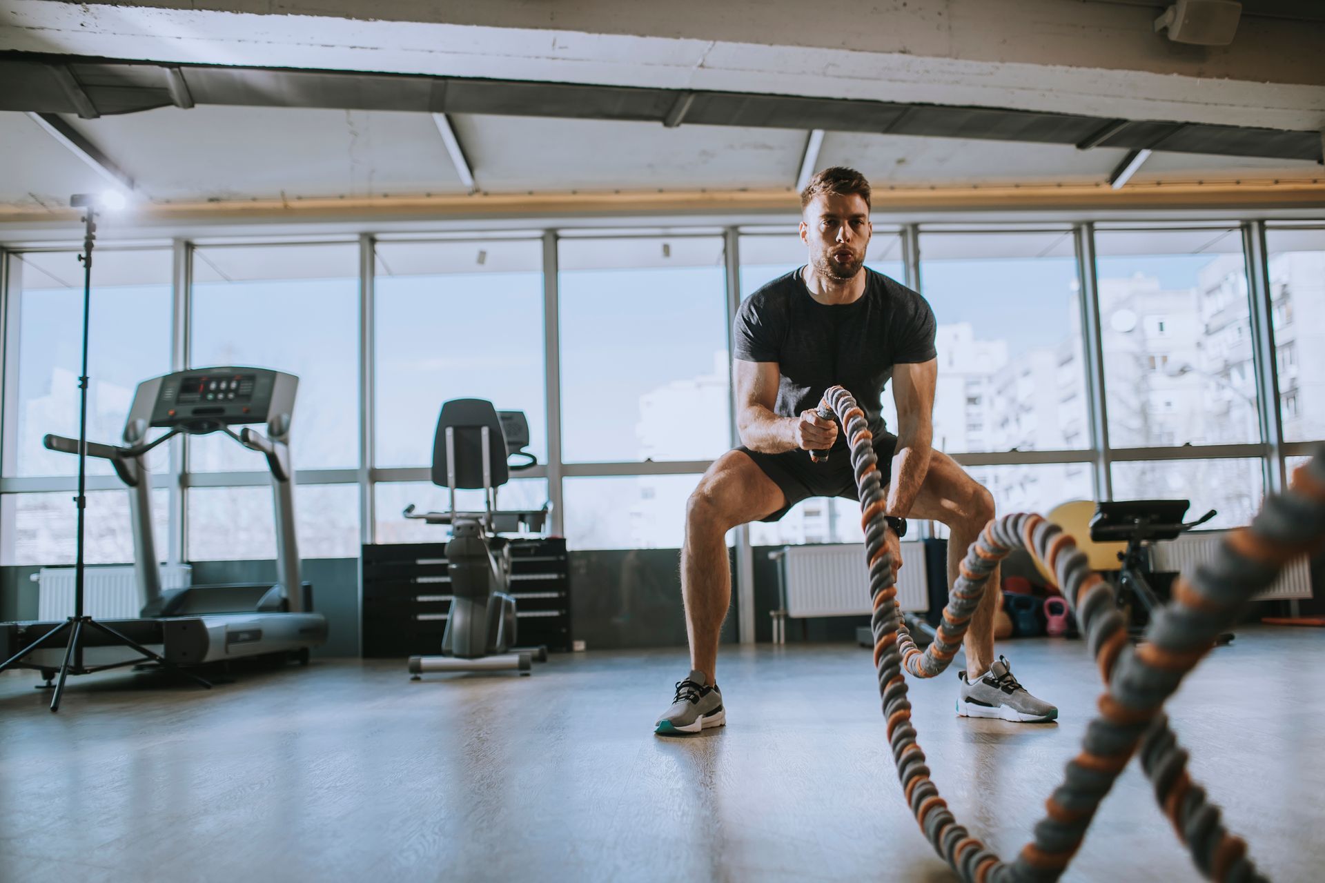 A person in a gym performing a battle rope exercise in a wide stance, with large windows in the background.