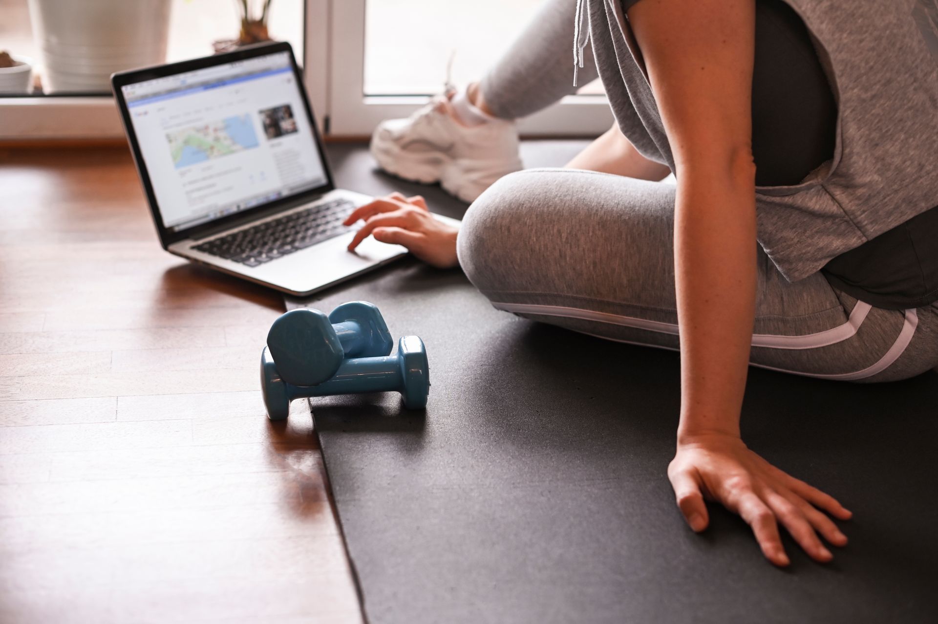A person sits on a yoga mat with blue dumbbells, using a laptop to guide their home workout.