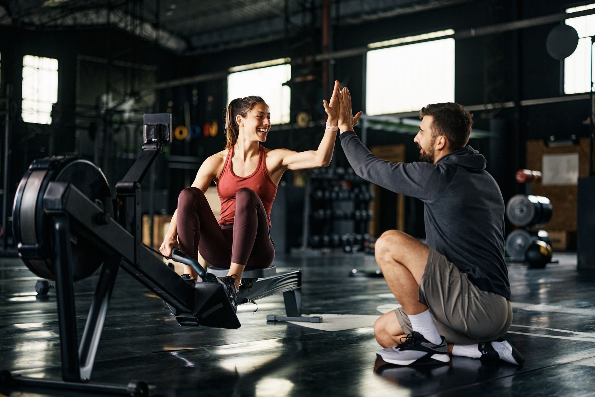 A person on a rowing machine in a gym high-fives a trainer who is kneeling beside them.