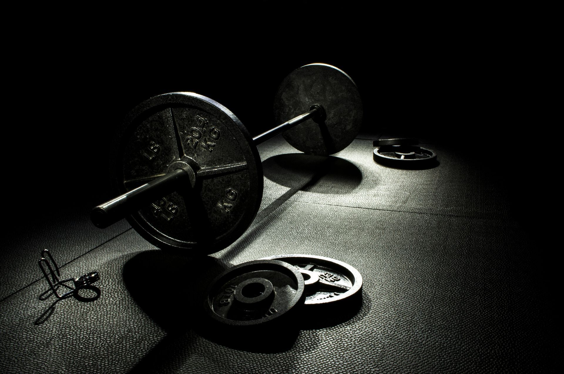 A low-angle, dark-lit shot of a barbell with weight plates resting on a gym floor, with extra weight plates nearby.
