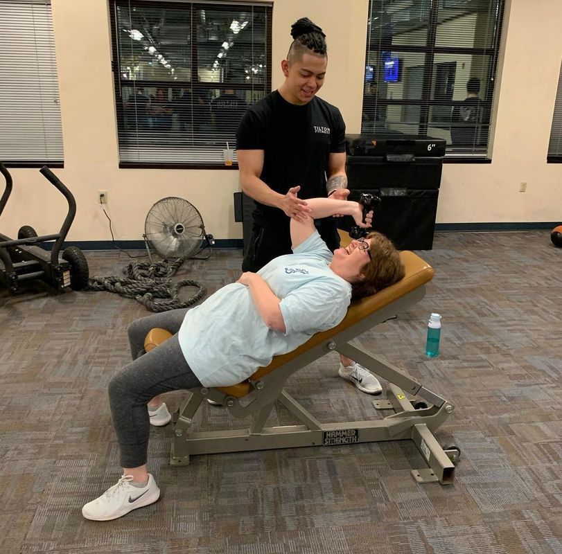 A fitness trainer assists a person lying on an inclined weight bench while they perform a dumbbell exercise in a gym.