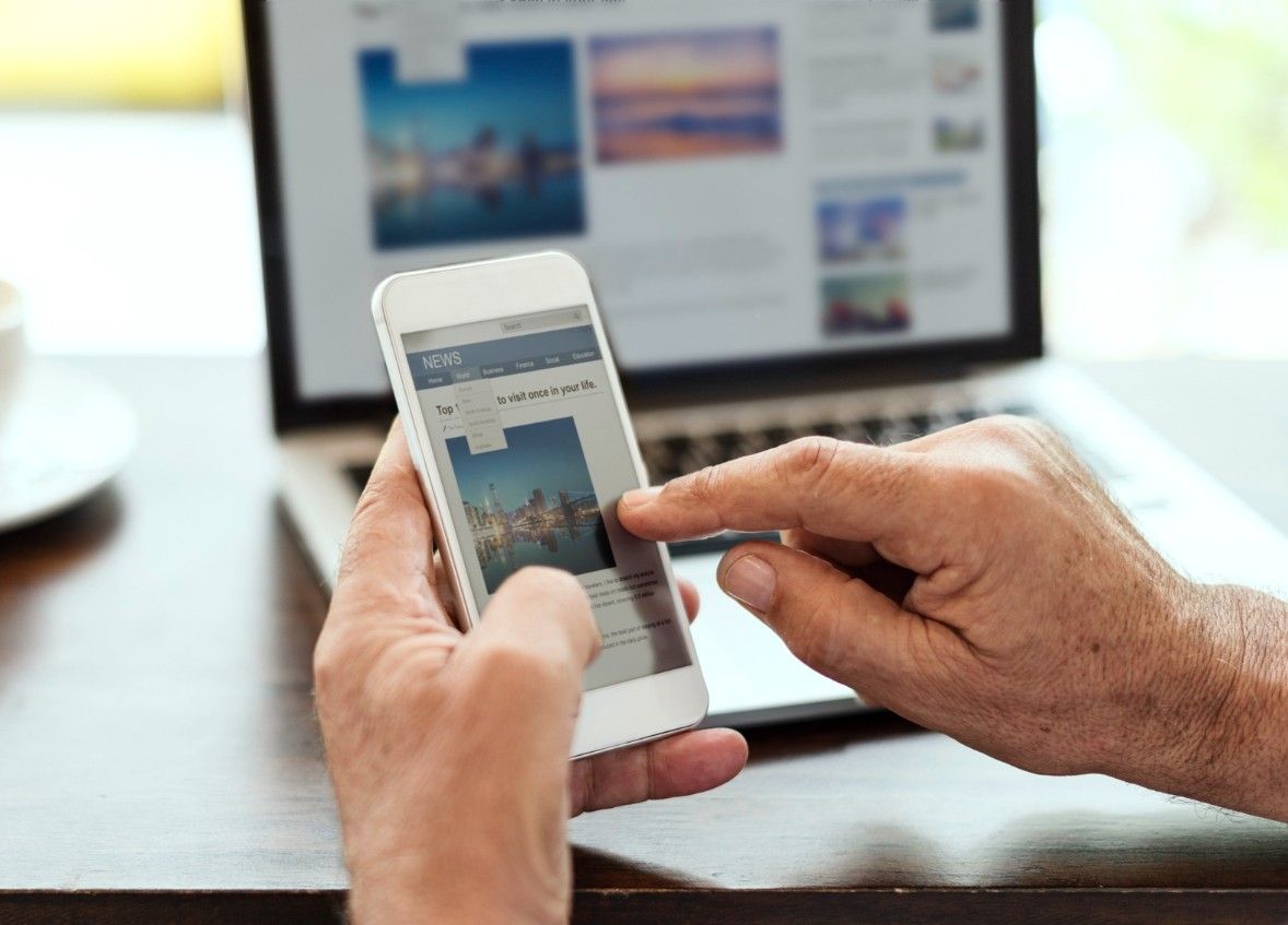 Person reading mobile-friendly content on a smartphone, with a laptop open in the background at a ca