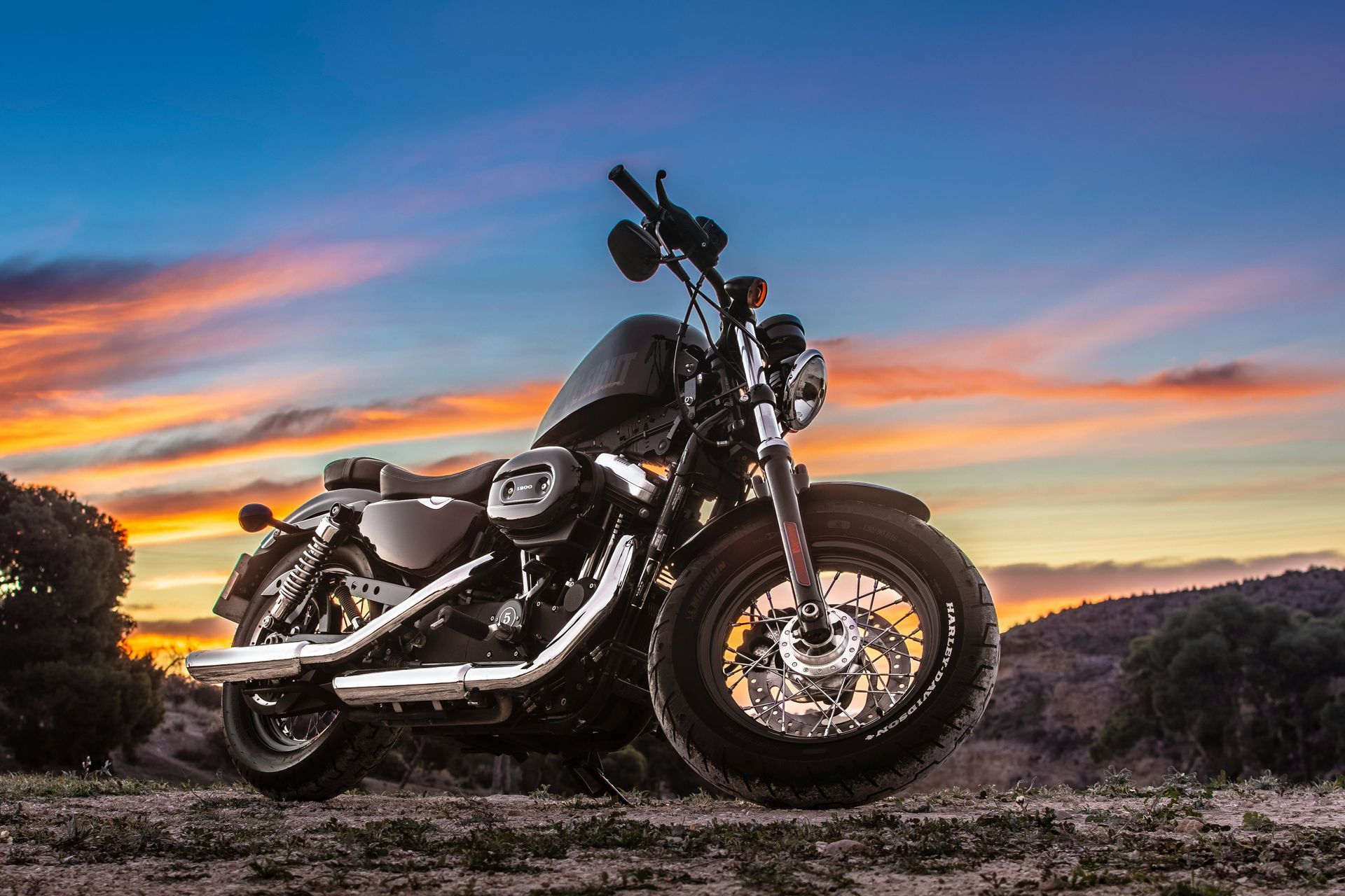 Black motorcycle parked on gravel at sunset with a colorful sky and rolling hills in the background