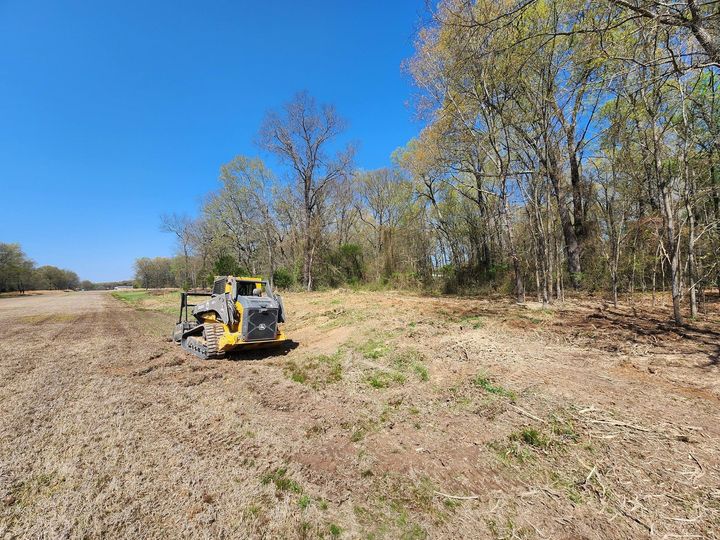 Yellow bulldozer clearing a dry field beside trees under a blue sky
