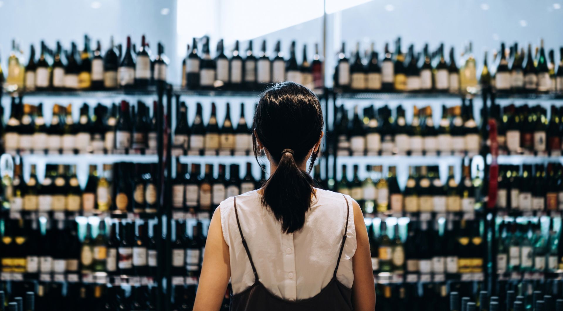 Rear view of woman grocery shopping for wines in supermarket liquor aisle, unsure which wine to choo