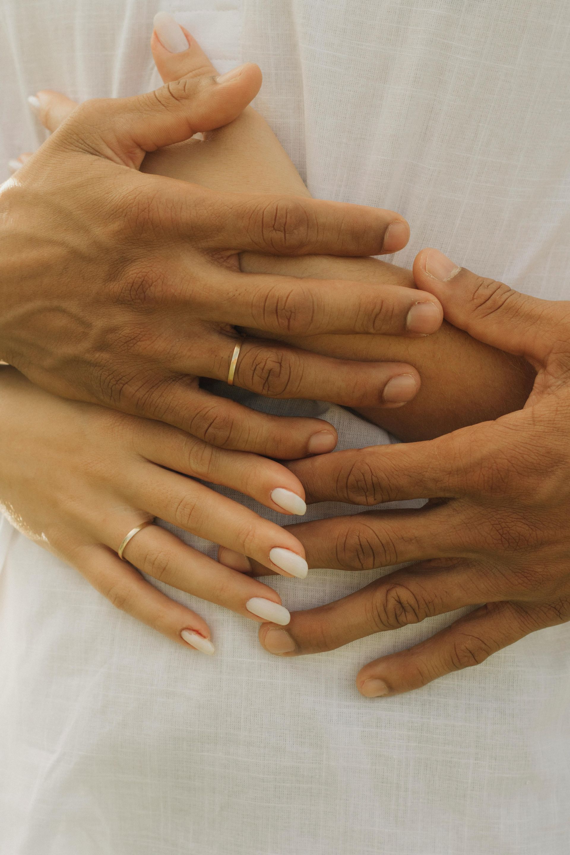Two sets of hands, one lighter-skinned, one darker, intertwined, wearing gold rings, set against a white fabric.