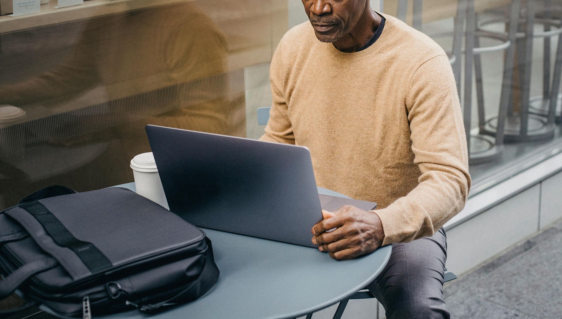 An older Black man in a tan sweater working on a laptop at an outdoor cafe.