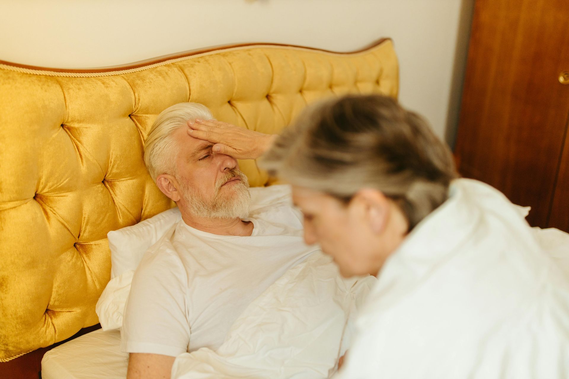 Older man in bed with a fever, being cared for by his wife.