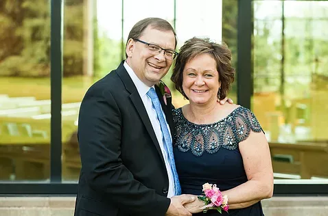 Man and woman smiling, formal attire, man in suit with blue tie, woman in navy dress, outdoors.