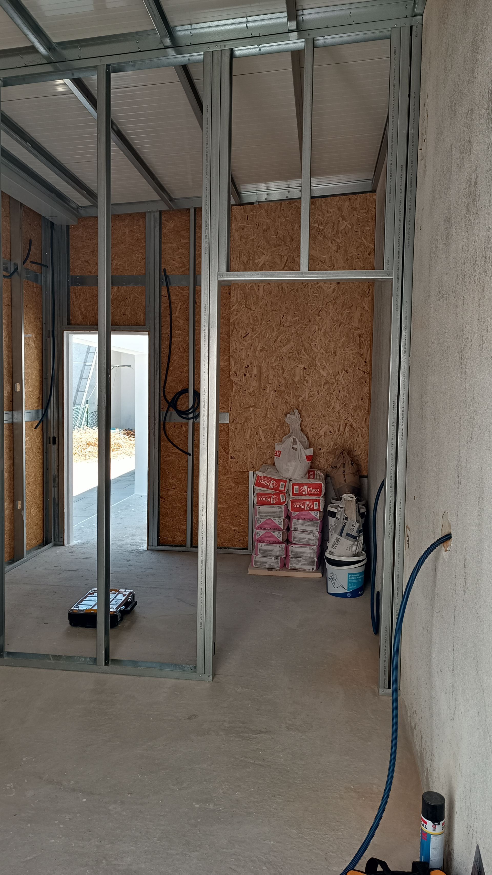 Interior de um cômodo em construção com estrutura metálica e paredes com material aparente. A porta dá acesso ao exterior.