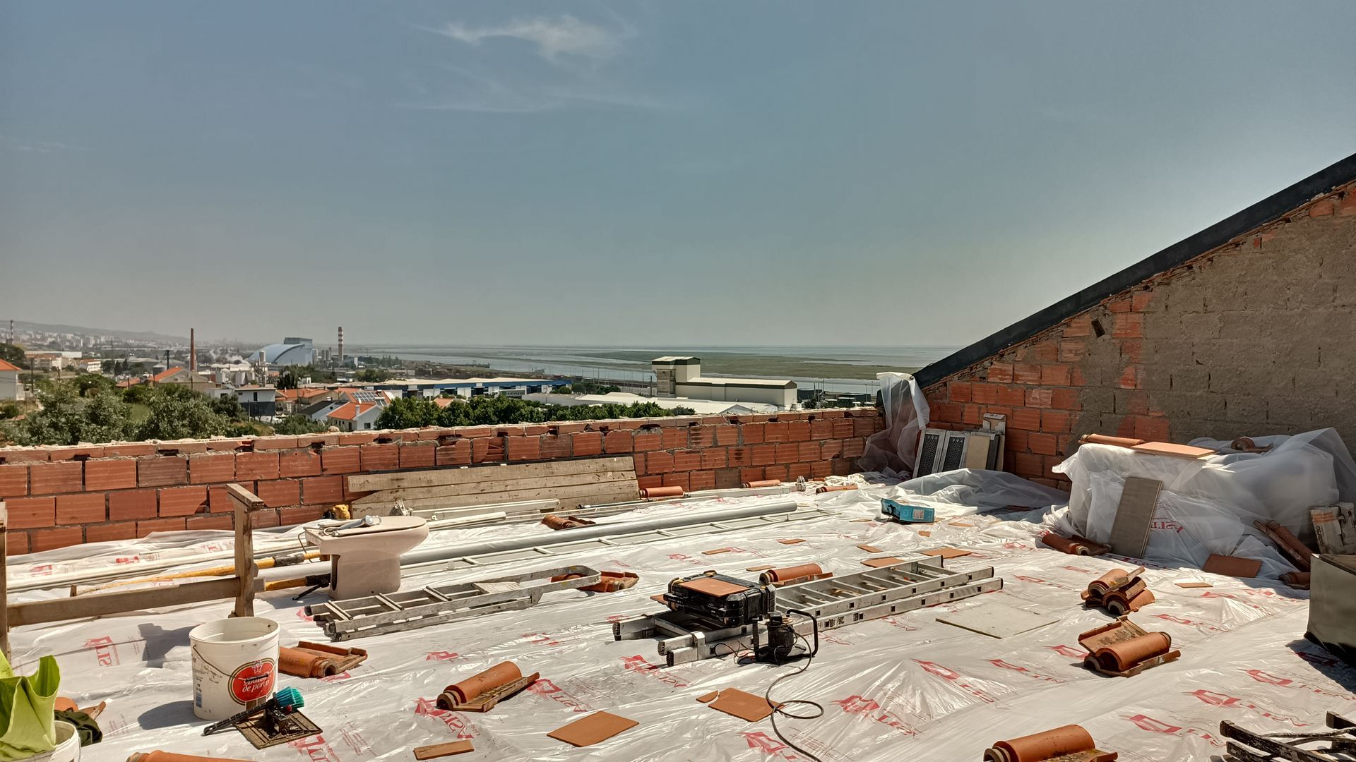 Telhado em construção com telhas de terracota. Paredes de tijolos, céu limpo, paisagem urbana ao fundo.