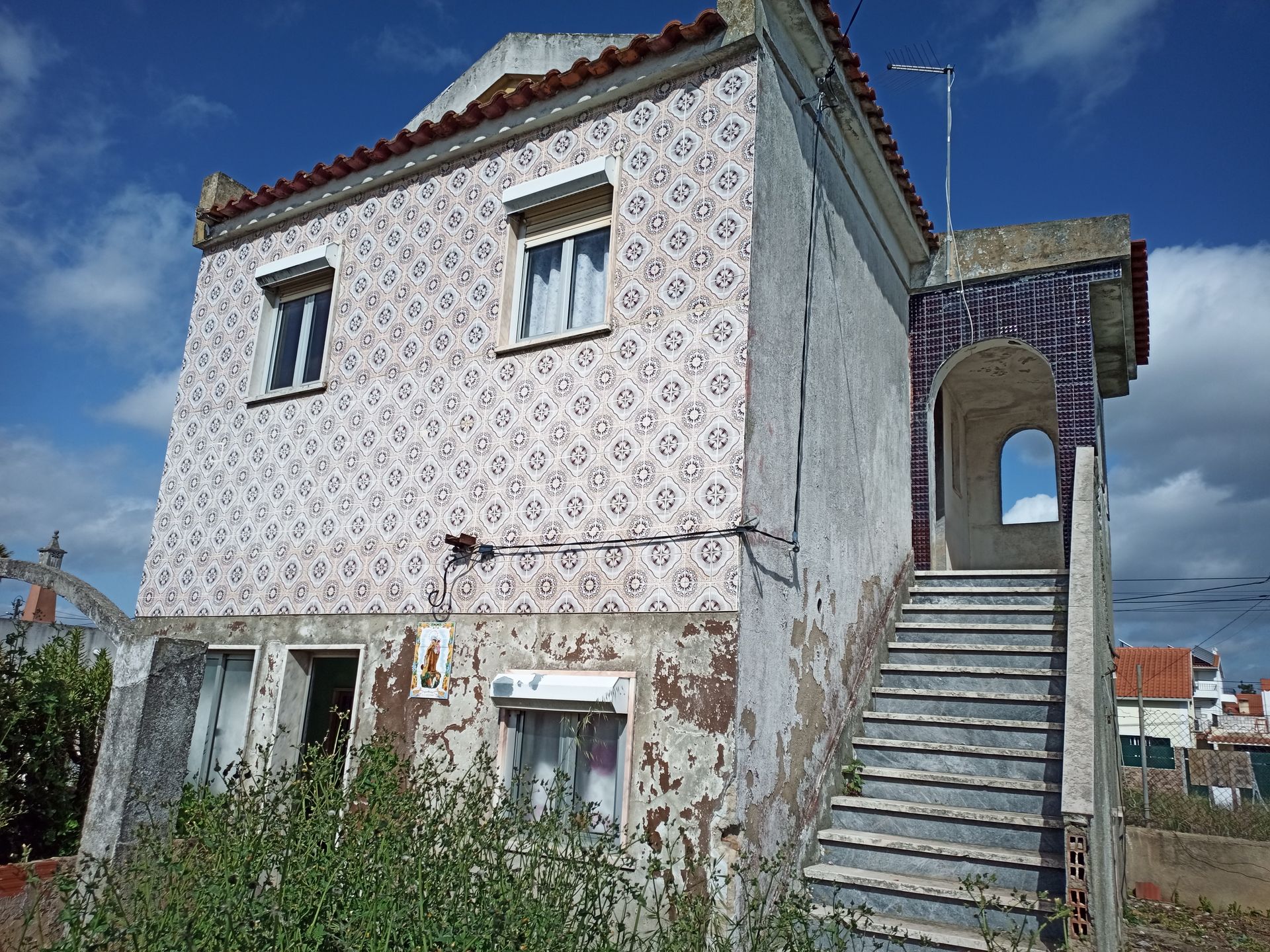 Edifício de dois andares com fachada revestida de azulejos estampados, escadas externas e paredes inferiores parcialmente expostas.