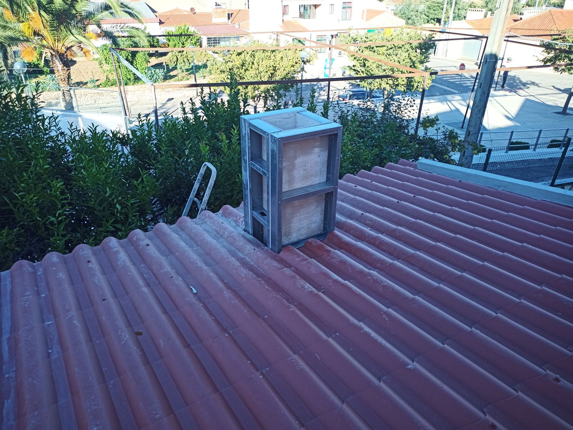 Uma chaminé de metal e tijolo se ergue sobre um telhado vermelho de zinco, cercada por arbustos verdes.