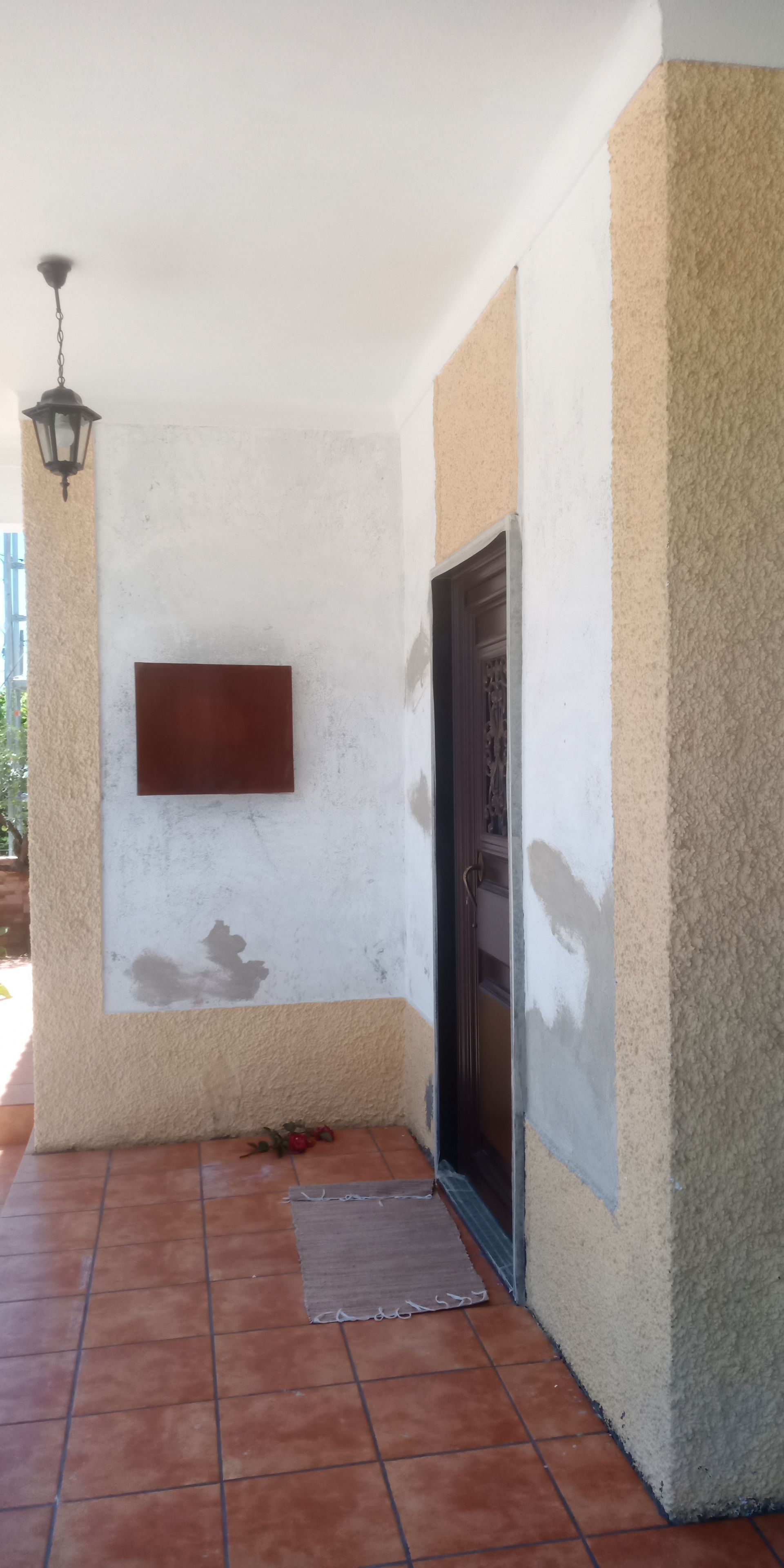 Varanda coberta com paredes de estuque descascando, porta marrom, piso de ladrilho de terracota e uma lanterna suspensa.