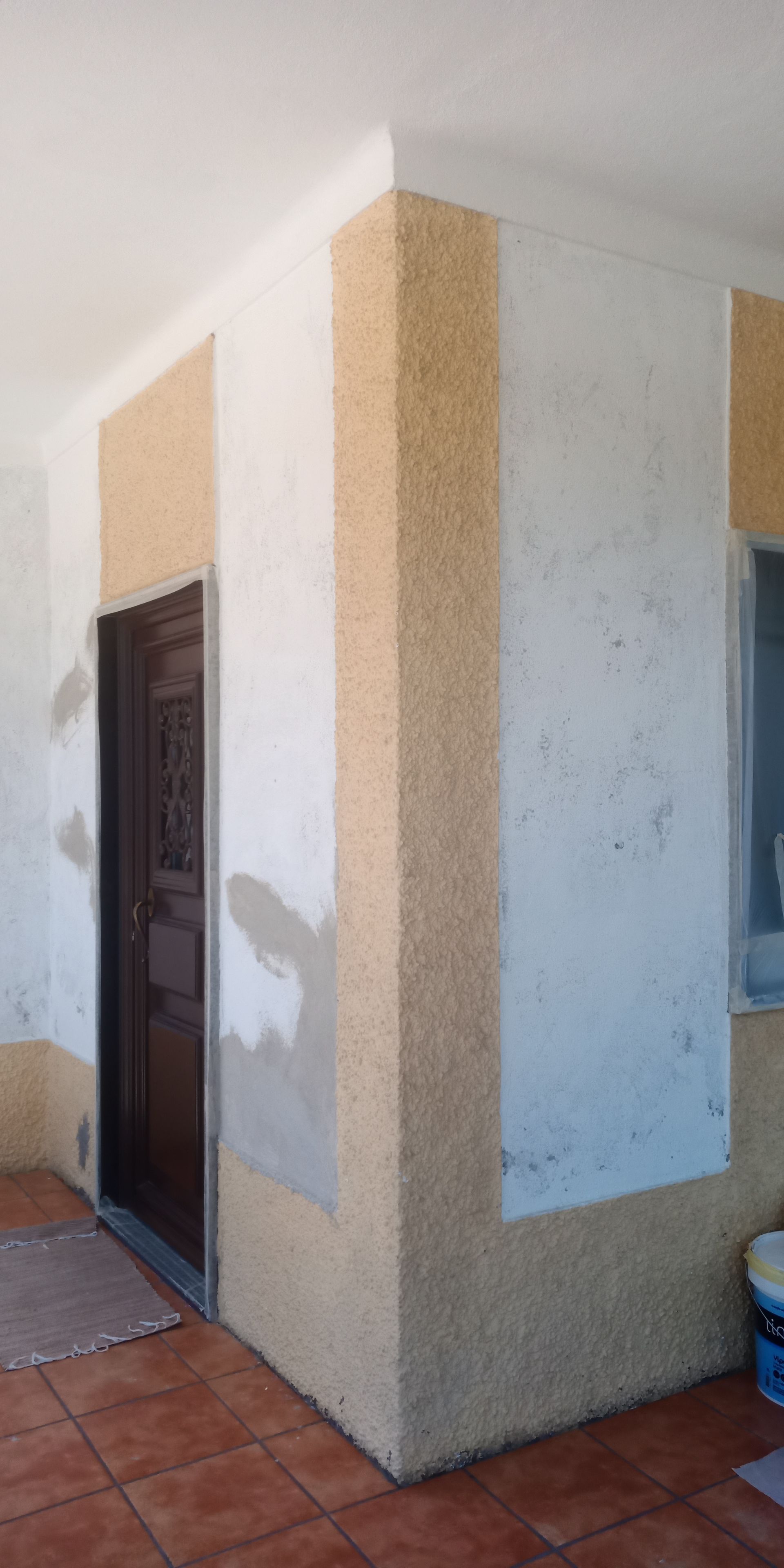 Canto exterior de um edifício, com porta. Paredes bege e brancas, piso de ladrilhos marrons.