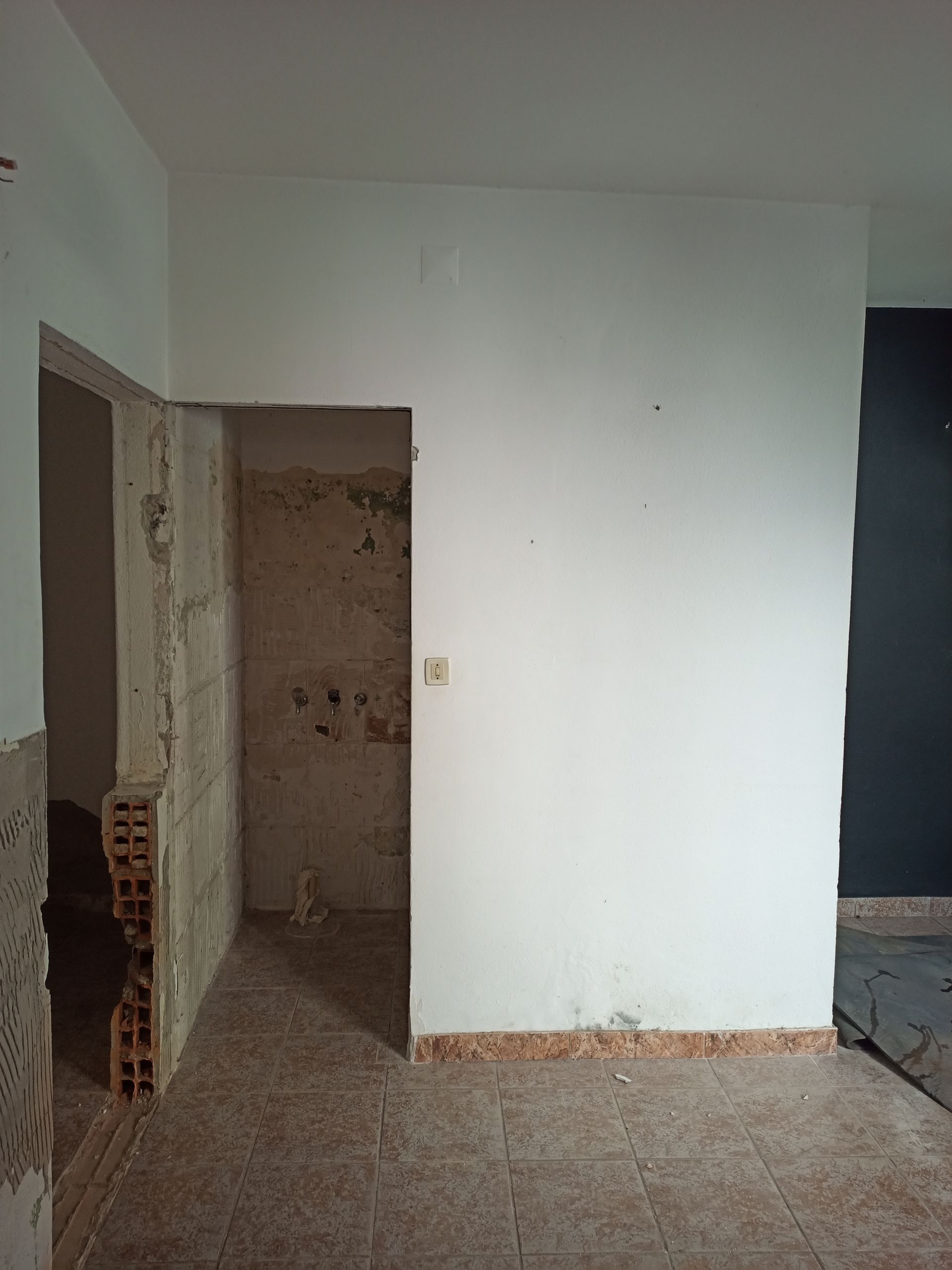 Vista interior de um cômodo em reforma, com porta e tijolos aparentes. Paredes brancas e piso de azulejo.